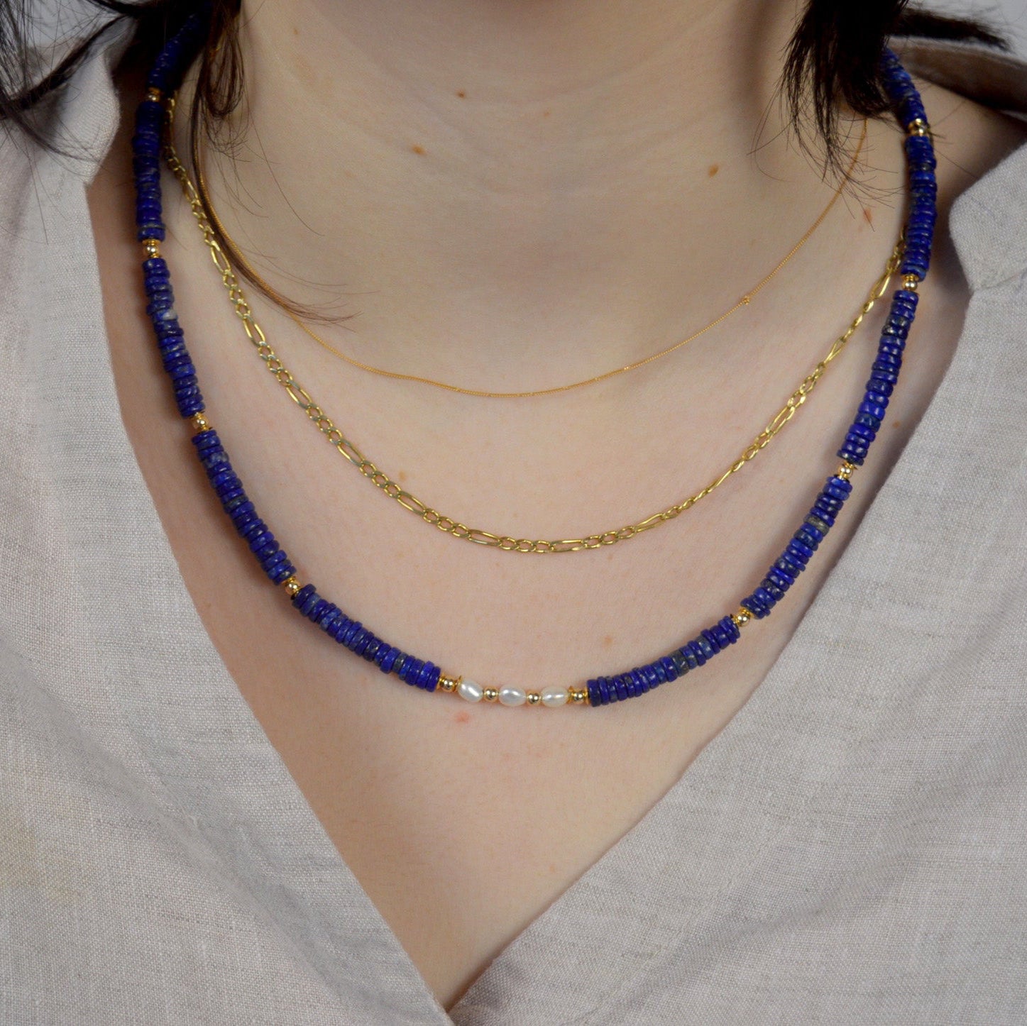 Two necklaces, one blue beaded and one gold chain, on a person wearing a light gray garment.