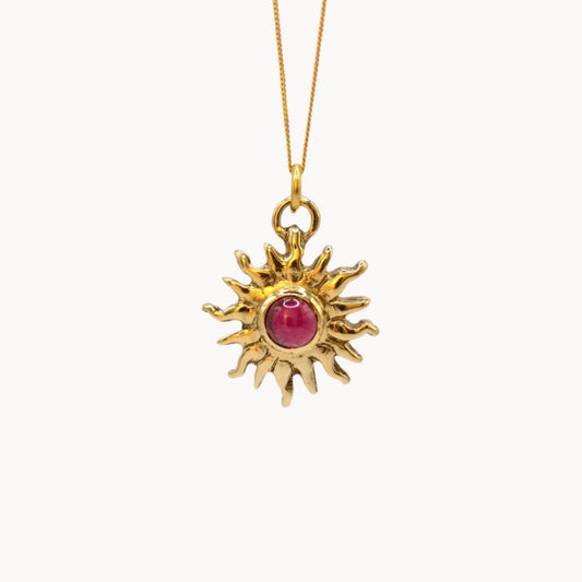 Gold sun-shaped pendant with a red gemstone on a white background